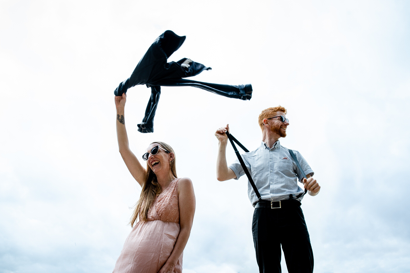 Happy wedding couple laughing outdoors holding jackets in the air under bright sky during relaxed countryside wedding portrait