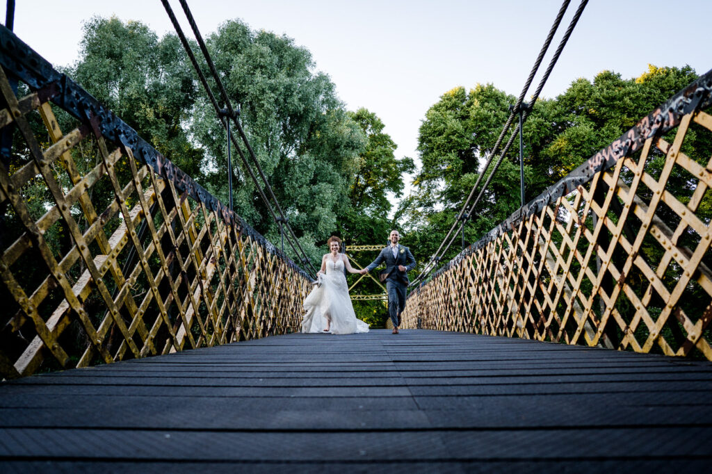 bride and groom run across a bridge holding hands towards the photographer