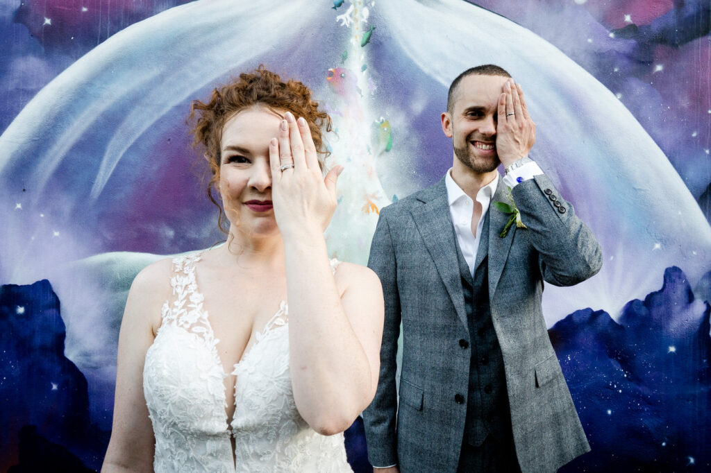 a couple stand in front of graffiti with their hands over their eyes showing the rings
