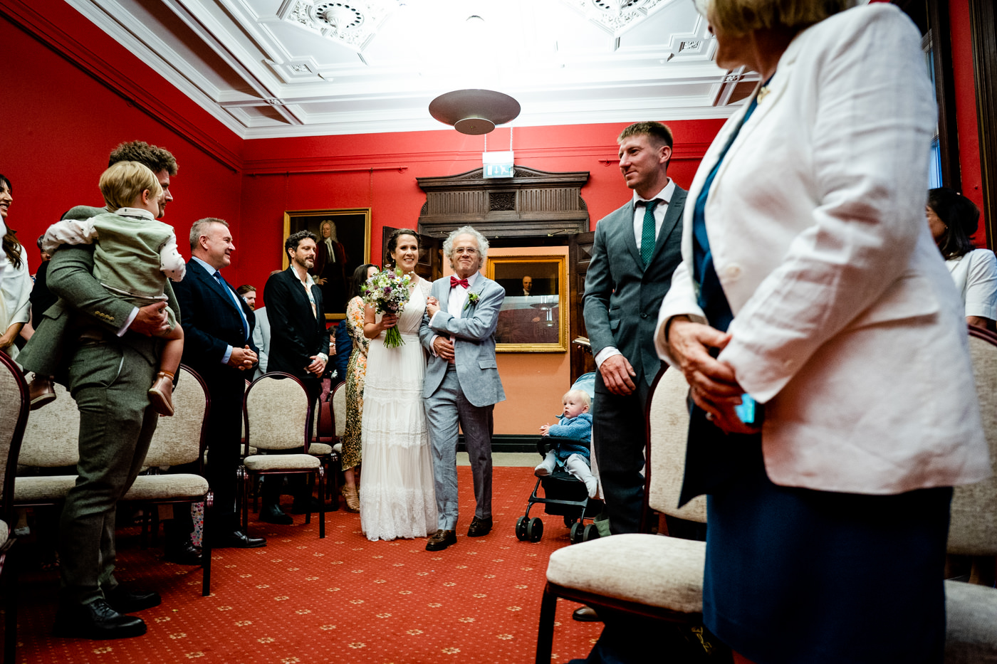 Bride walking into the Bristol Registry Office wedding ceremony holding a bouquet with family beside her.