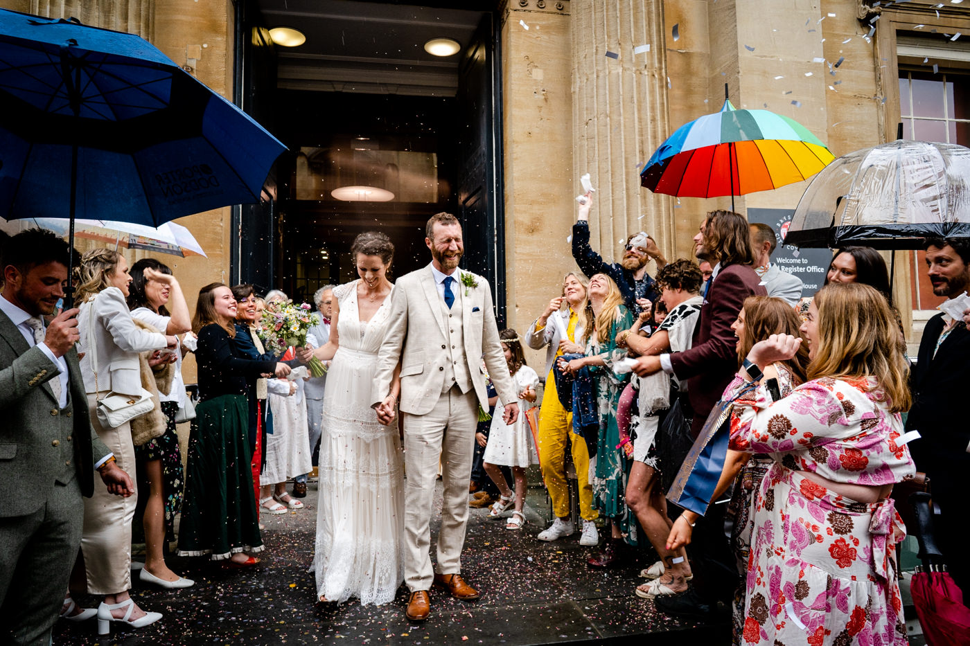 Couple walking through confetti outside Bristol Registry Office while guests hold colourful umbrellas in the rain.