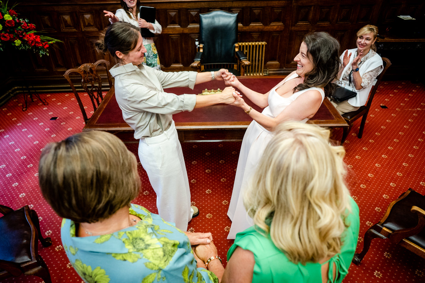 Two brides holding hands and exchanging rings at a table during their Bristol Registry Office wedding ceremony.